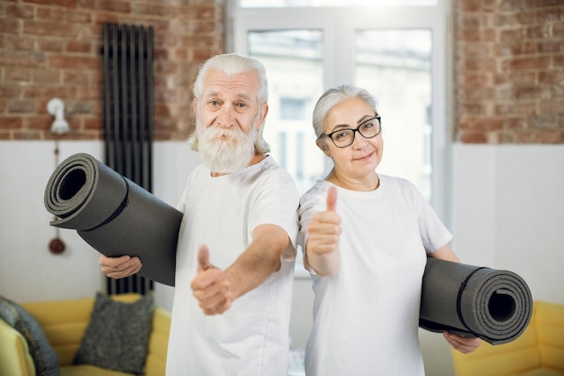 Positive aged couple showing thumb up and holding yoga mat