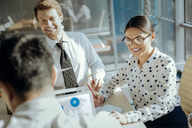 Positive ambience. Charming cheerful young woman and her male colleague sitting in their boss office and listening to his explanations while smiling at him