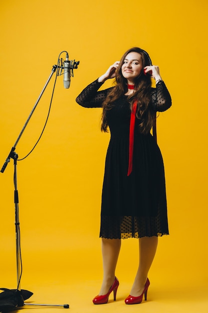 The positive amusing brunette female singer having fun sings\
song young female caucasian pop artist in earphones smiling while\
performing joyful song in microphone on the yellow background