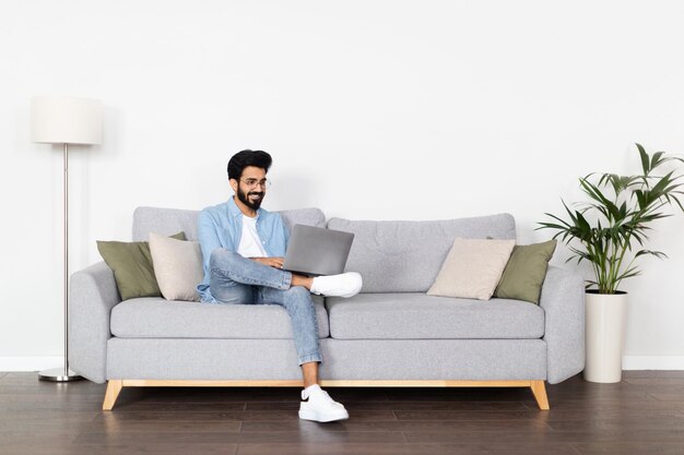 Positive arab guy using laptop sitting on couch at home