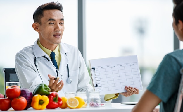 긍정적인 아시아 의사가 병원에서 건강한 음식을 들고 책상에 앉아 있는 동안 남성 환자에게 테이블을 보여주고 웃고 있다
