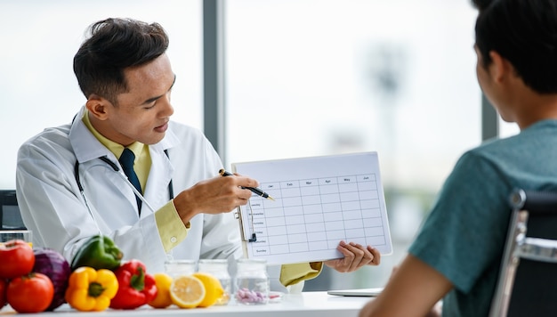 positive asian doctor smiling and demonstrating table to male patient while sitting at desk with healthy food in clinic