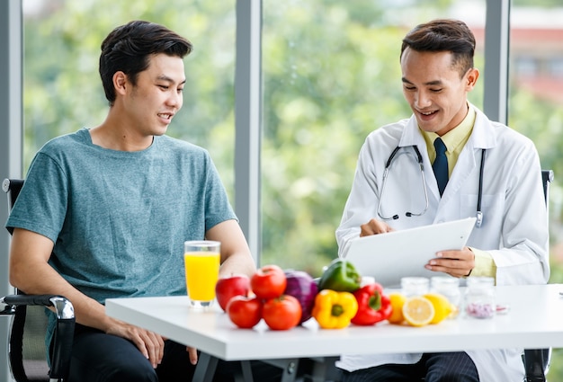 Positive Asian doctor smiling and demonstrating table to male patient while sitting at desk with healthy food in clinic