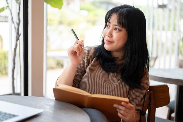 긍정적 인 아시아 여성 이 카페 에 앉아 일기 를 쓰거나 책 에 자신의 생각 을 나열 하고 있다