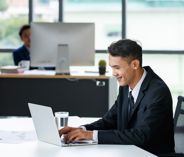 Imprenditore maschio asiatico positivo seduto al tavolo e navigando nel netbook mentre si lavora al progetto sul posto di lavoro e guardando lo schermo del computer portatile.