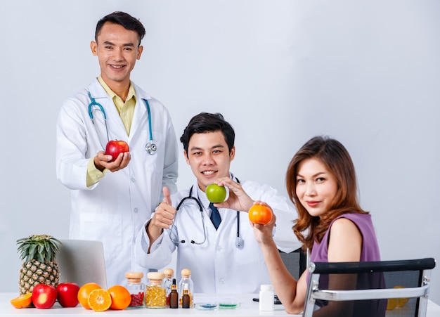 Positive Asian male nutritionists with fruits and healthy food gathering at table with female patient while looking at camera on white background