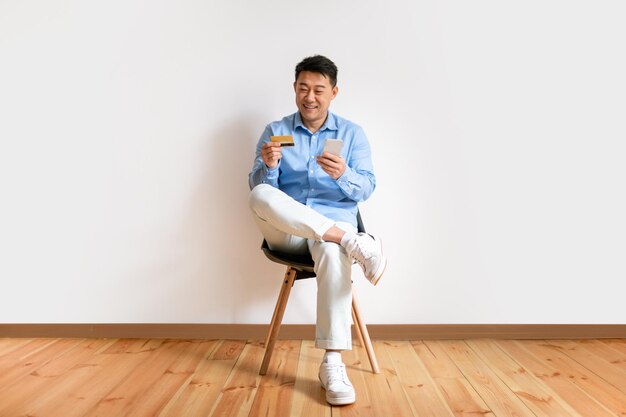 Позитивный азиатский мужчина делает покупки в Интернете с мобильным телефоном и кредитной картой, сидя в кресле против белого