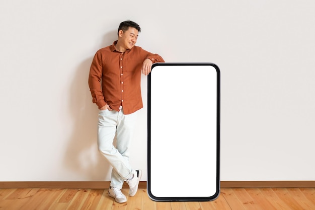 Positive asian mature man leaning on big cellphone with white screen template panel standing over white wall mockup