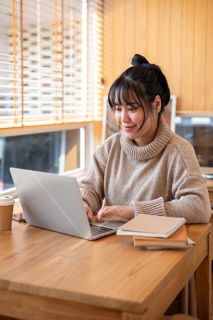 A positive Asian woman in a cosy sweater is working on her laptop computer in a cafe or library