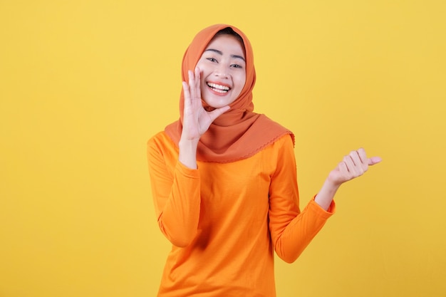 Positive asian woman girl points thumb, demonstrates copy space on blank yellow wall, has happy friendly expression, dressed casually wearing hijab, poses indoor