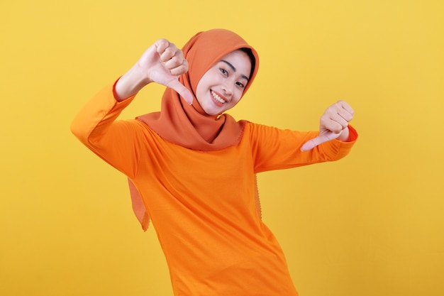 Positive asian woman girl points thumb down, demonstrates copy space on blank yellow wall, has happy friendly expression, dressed casually wearing hijab, poses indoor