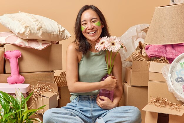 Positive asian woman holds vase of gerberas applies green\
hydrogel patches under eyes to reduce puffiness sits crossed legs\
around cardboard boxes against beige background relocation\
concept