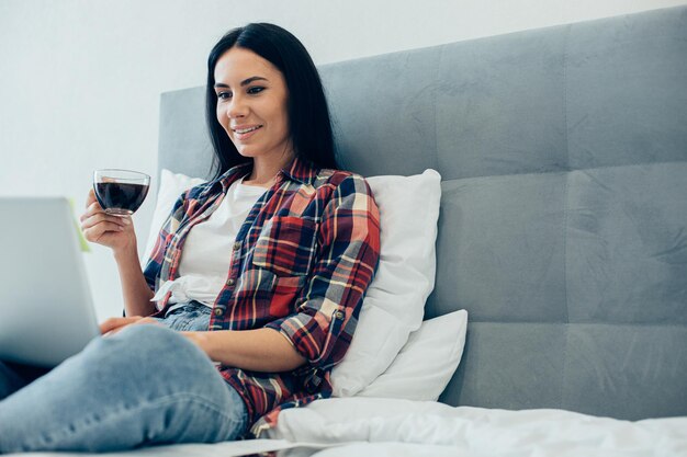 Positive attractive lady holding a cup of tasty coffee and looking at the screen of a laptop while sitting on the bed