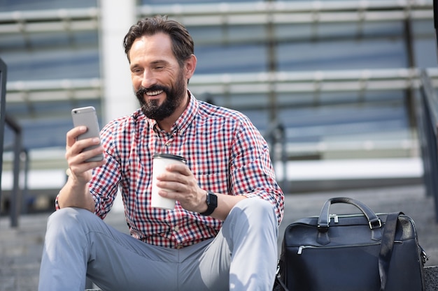 Positive bearded adult man sitting in urban settings while drinking coffee and using phone