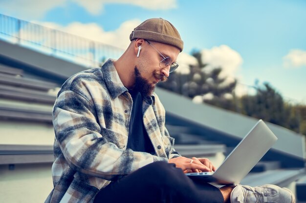 Positive bearded hipster guy sitting in outdoors and typing text on laptop g internet connection