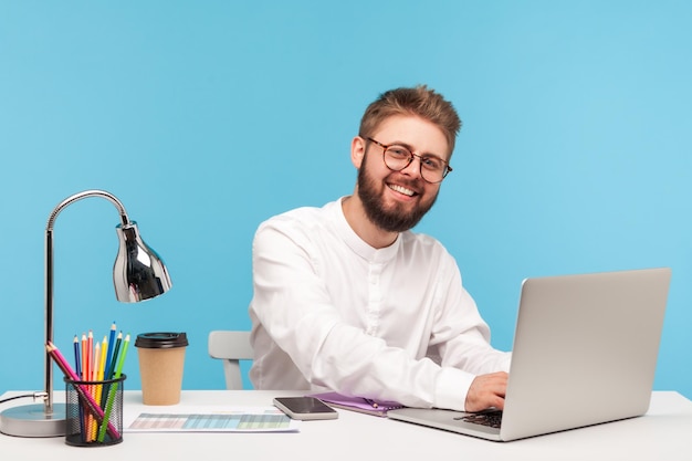 Positive bearded man office worker in eyeglasses and white shirt sitting at workplace typing on laptop and smiling looking at camera success Indoor studio shot isolated on blue background