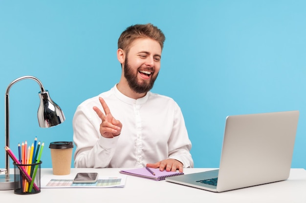 Позитивный бородатый офисный работник, показывающий V-жест пальцами, говорящий по видеозвонку, смотрящий в камеру ноутбука с зубастой улыбкой, сидящий на рабочем месте. Крытая студия снята на синем фоне