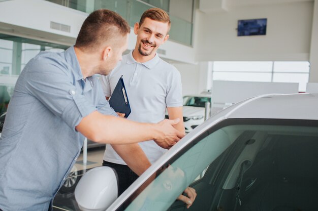Positive and bearded young seller stand in front of customer and looks at him. He is smiling. Buyer touches car and looks at seller. He is talking seriously.
