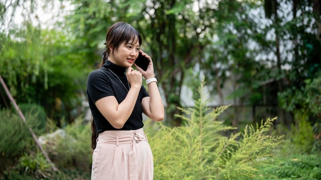 Photo a positive beautiful asian woman talking on the phone while walking in a beautiful green garden