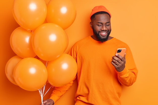 Positive birthday guy receives congratulation messages on smartphone celebrates anniversary dressed in sweater and hat poses with balloons waits for guests to come on party isolated over orange wall