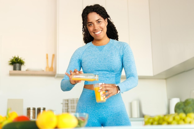 Positive black fit woman enjoying tasty lunch and pouring orange juice from jug to glass standing in kitchen at home