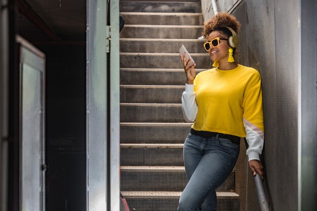 Позитивная черная хипстерская женщина в модном наряде с помощью мобильного телефона