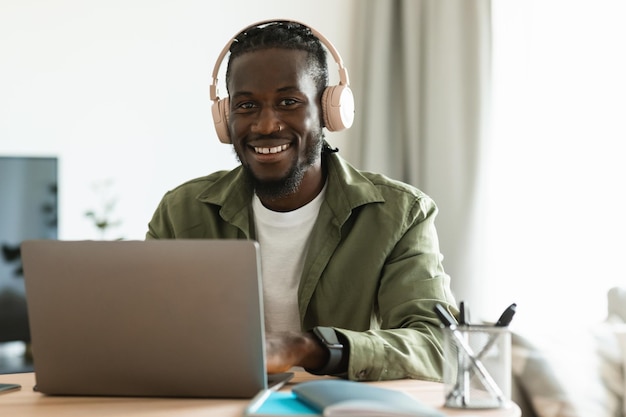 Positive black man in wireless headphones working online on laptop at home office looking and smiling at camera