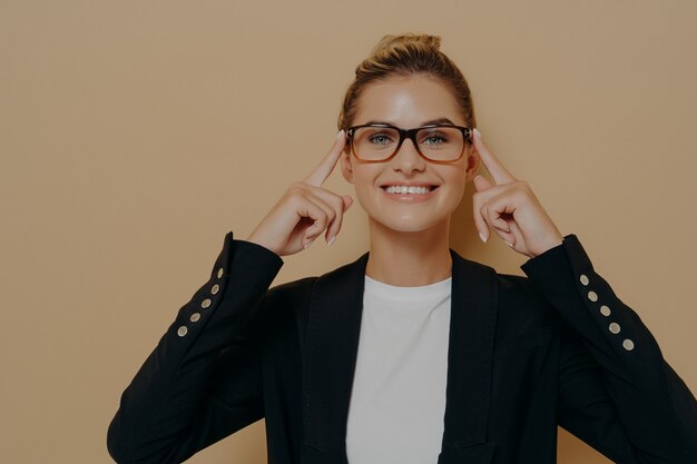 Positive blonde female student putting on eyeglasses and holding them with hands, young beautiful caucasian woman posing and looking through spectacles at camera with beaming smile. Eyesight concept