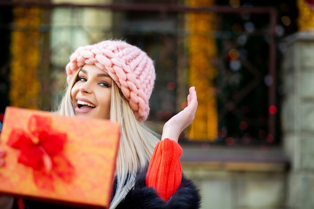 Positive blonde girl wearing fur coat and cap pulls a gift box to you. Space for text