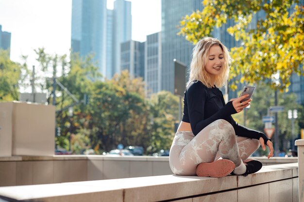 Позитивная блондинка использует мобильный телефон на открытом воздухе Спортсменка в спортивной одежде отдыхает после тренировки на улице большого города Небоскребы на заднем плане