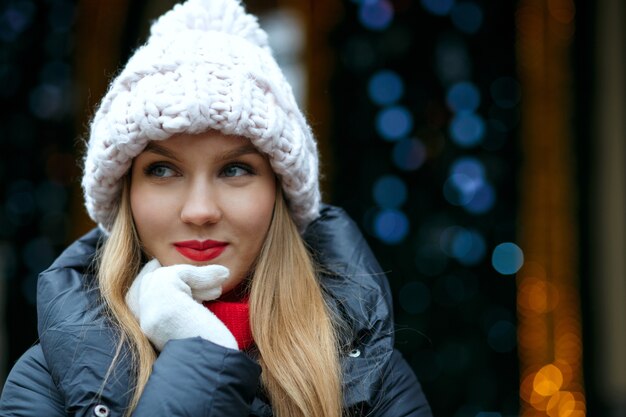 Positive blonde model with red lips enjoying winter holidays at the street over a garlands background