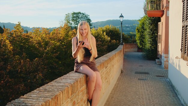 Positive blonde woman making video call with smartphone from the place of her visit. Summer time in other country.