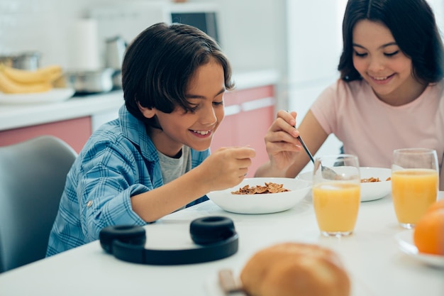 Positive boy and girl smiling while sitting at the table at home and eating cornflakes from the bowls