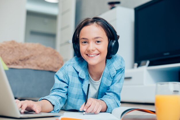 Positive boy wearing headphones and holding a pencil while lying on the floor in front of a laptop screen