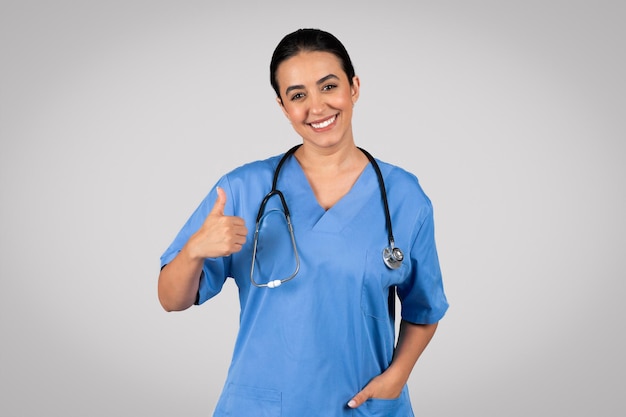 Positive brazilian woman doctor in uniform showing thumb up recommending medicine service gray