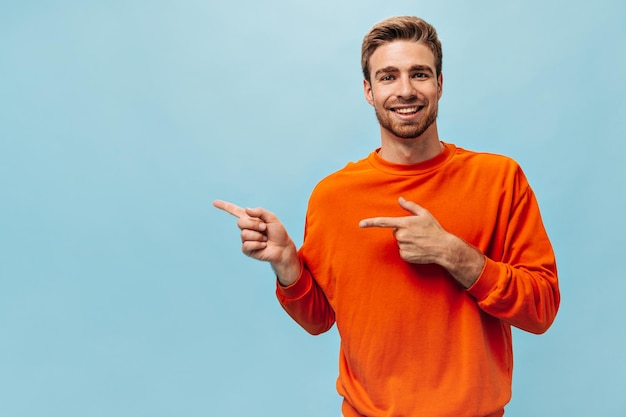 Positive bright guy with ginger beard and brown hair in orange cool clothes smiling and pointing to place for text on blue backdrop