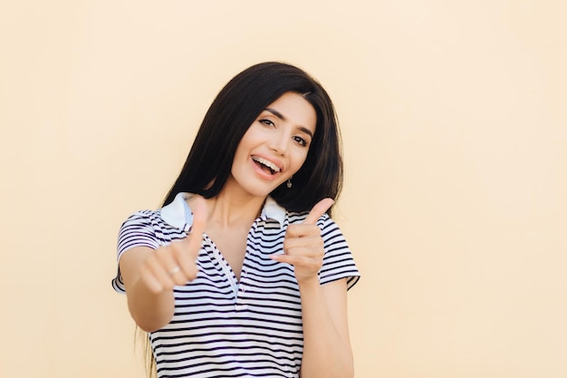 Positive brunette female makes approval gesture keeps thumbs\
raised being in good mood rejoices something isolated over light\
pink background optimistic woman agrees or evaluates new\
project