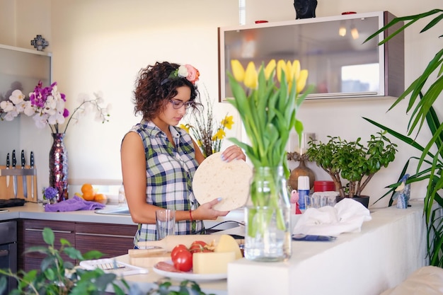 ポジティブなブルネットの女性は、背景に黄色いチューリップのあるキッチンで生地を作ります。