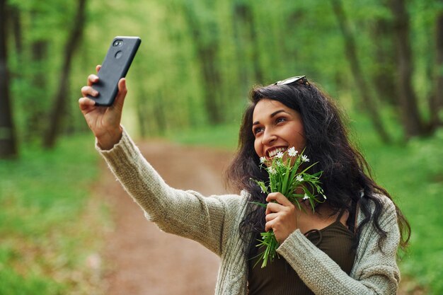 Positive brunette holding flowers and taking selfie by phone in the forest