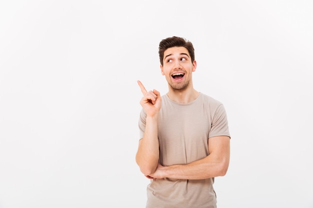 Positive brunette man 30s wearing beige t-shirt gesturing finger aside on copyspace, isolated over white wall