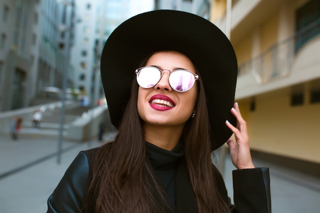 Positive brunette model in mirror glasses and broad brimmed hat posing near the city building