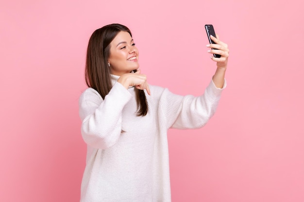 Positive brunette woman taking selfie or having livestream pointing finger down subscribe her vlog wearing white casual style sweater Indoor studio shot isolated on pink background