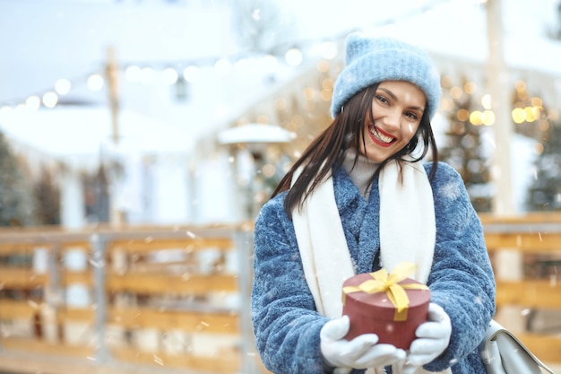 Positive brunette woman in winter coat holding a gift box at christmas fair during the snowfall. Space for text