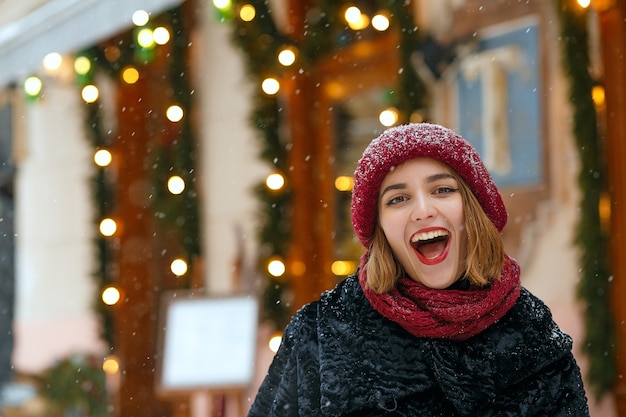 빨간 모자와 스카프를 두르고 눈이 내리는 동안 도시를 걷고 있는 붉은 입술을 가진 긍정적인 브루네트 여성. 빈 공간