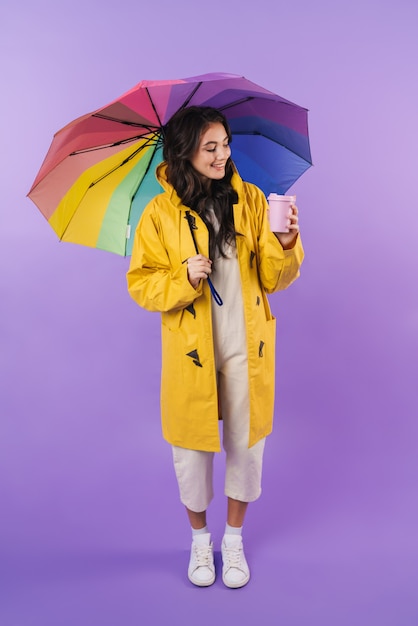 positive brunette woman in yellow raincoat posing isolated over purple wall wall holding umbrella and coffee.