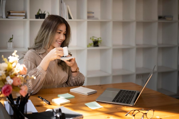 Donna d'affari positiva che beve caffè e legge notizie sul suo computer portatile