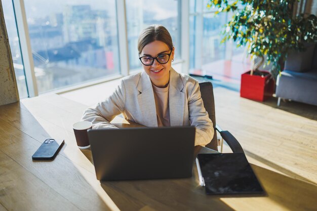 A positive business woman in a jacket is sitting at a table working on a laptop in a bright office A young European woman is sitting at a table Freelance and remote work Modern women's lifestyle