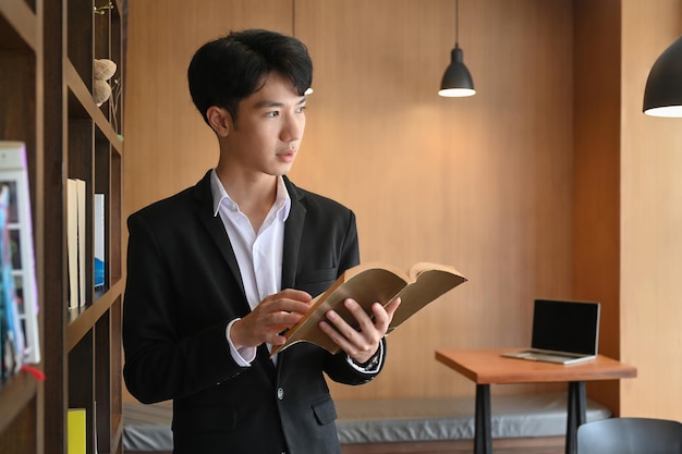 Positive businessman holding book while standing near bookshelf