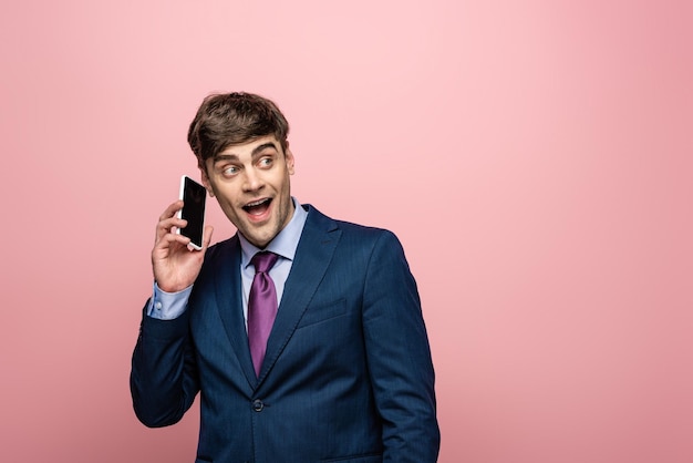 Позитивный бизнесмен разговаривает по смартфону и смотрит в сторону на розовом фоне