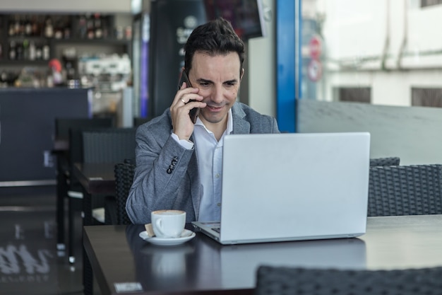 Uomo d'affari positivo con il computer portatile che parla sul telefono nella caffetteria